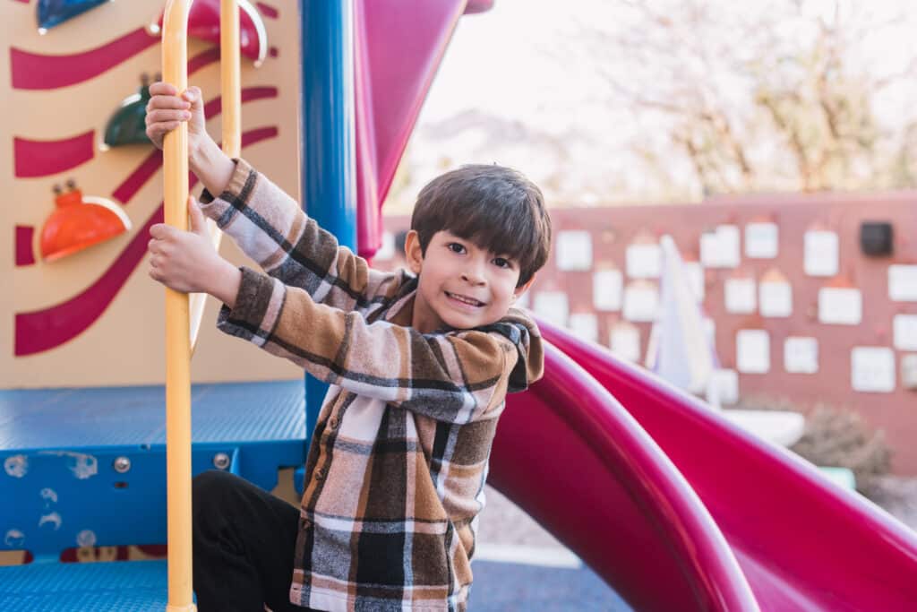 A young boy with a full head of dark brown hair plays on a fire pole in the colorful playground in the backyard of the Ronald McDonald House.