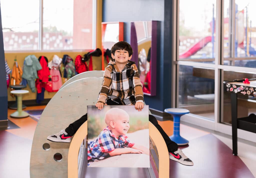 A young boy with dark brown hair and a plaid shirt poses with a canvas print. The canvas print is a photo of him from 3 years ago, after he lost his hair from chemotherapy.